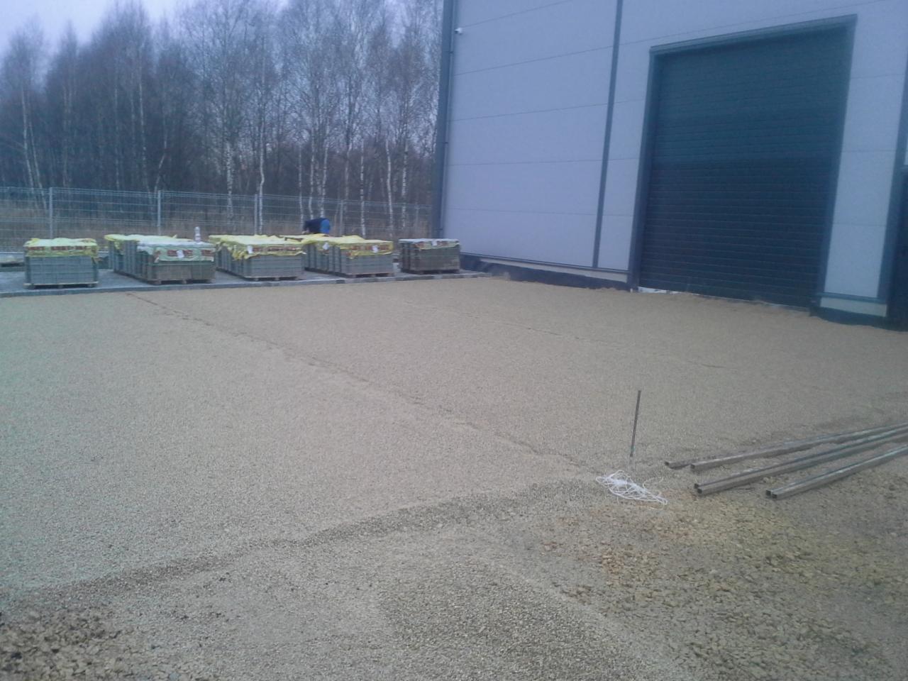 Przygotowanie podłoża pod kostkę: warstwa podsypki z kamyczków, rury i sznurki do poziomowania, palety z kostką brukową w tle przy hali.