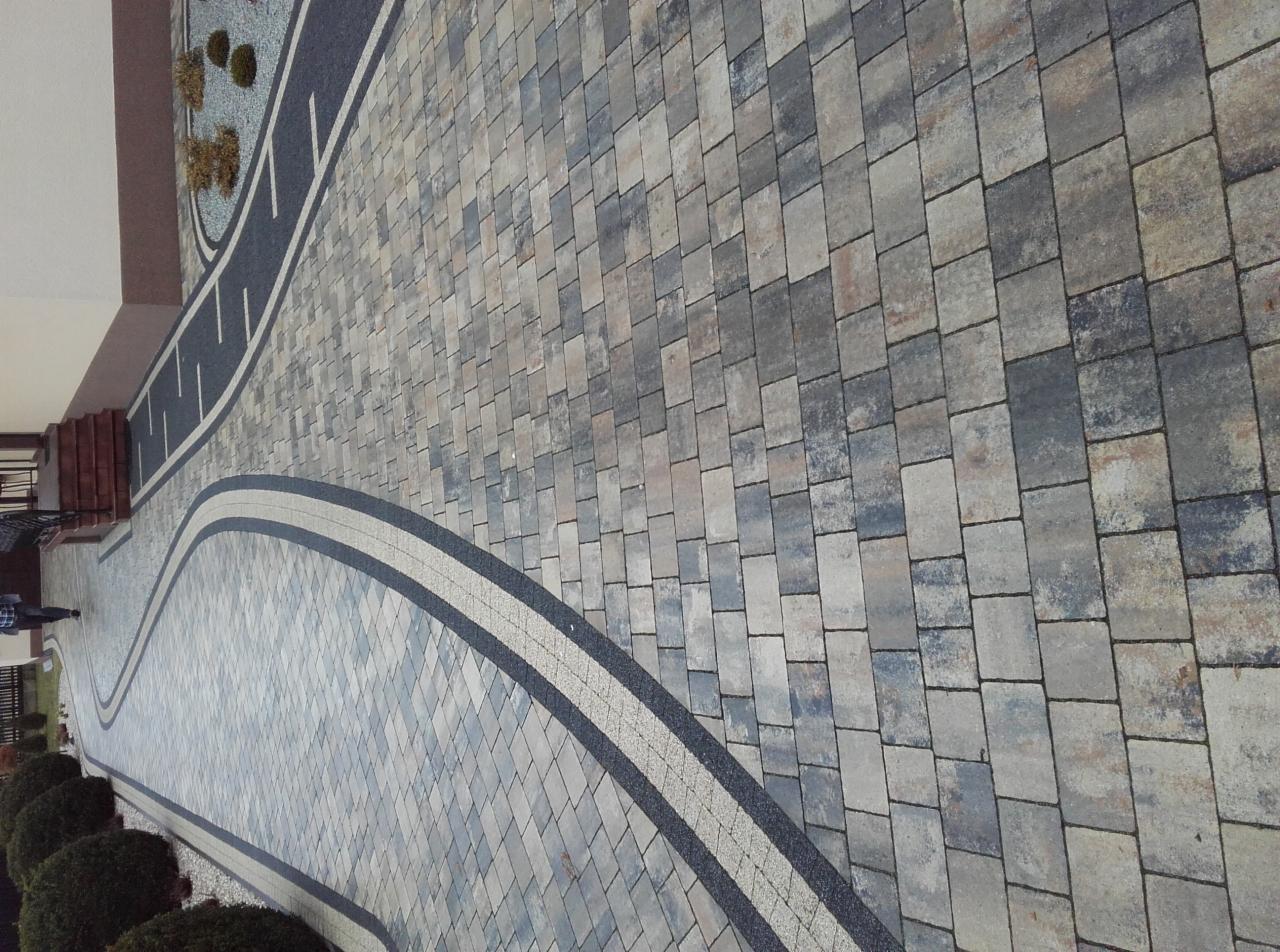 Wzorzysta, nowa nawierzchnia z kostki brukowej w odcieniach szarości i beżu, z dekoracyjnymi pasami obrzeży. Widok z góry, na pierwszym planie roślinność.