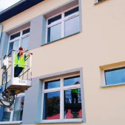 Pracownik w zielonej kamizelce na podnośniku koszowym, myjący okna na wysokości w budynku z szarymi ramami okiennymi, widoczne odbicie w oknach.
