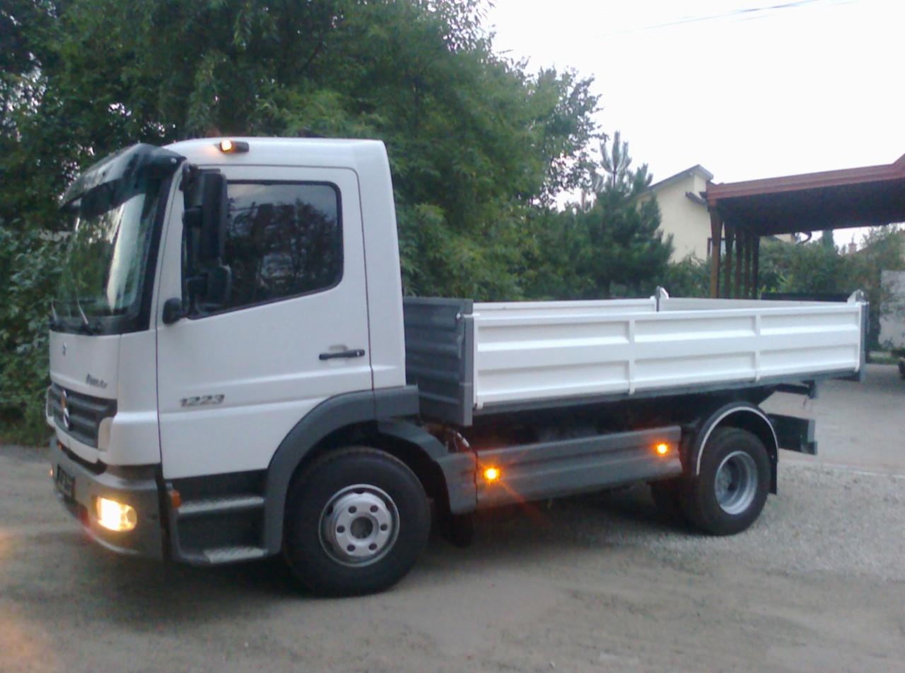 Biała ciężarówka Mercedes-Benz 1223 z otwartą skrzynią ładunkową, zaparkowana na żwirowym podłożu, z zielenią w tle. Widoczne światła pozycyjne.