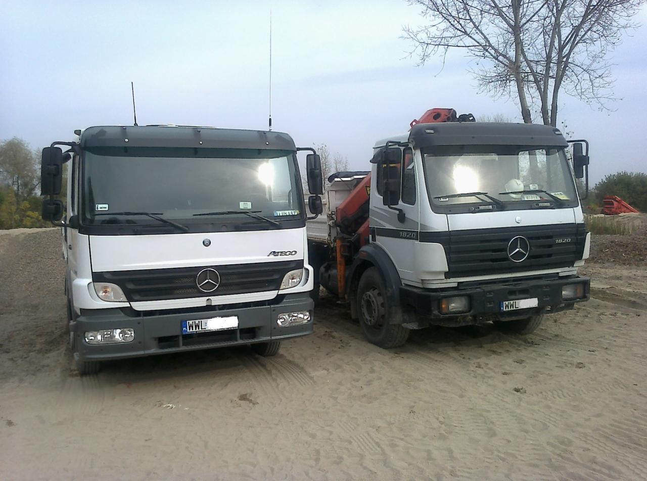 Dwa samochody ciężarowe Mercedes: Atego i 1820, stoją obok siebie na piaszczystym terenie. Atego po lewej stronie, 1820 z dźwigiem po prawej. Widoczne tablice rejestracyjne.