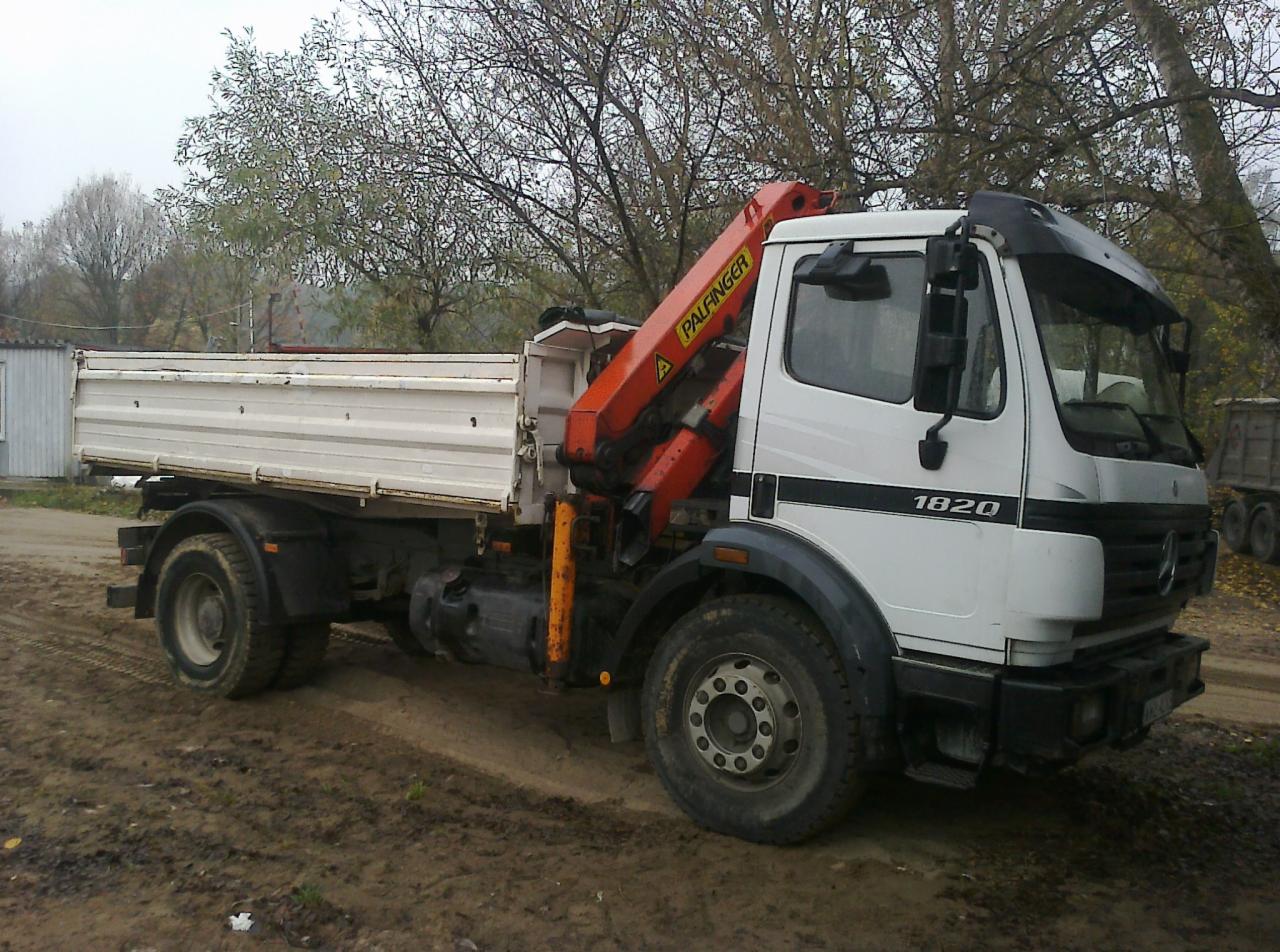 Biała ciężarówka Mercedes-Benz 1820 z pomarańczowym dźwigiem Palfinger na błotnistym terenie. Widoczne drzewa w tle, pochmurne niebo.