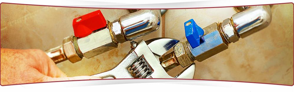 Regulowany klucz nastawny dokręca złączkę rury z niebieskim zaworem na tle beżowych płytek. Widoczna dłoń hydraulika.