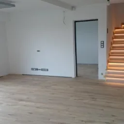 Jasne panele podłogowe w przestronnym wnętrzu z oknem, schodami z podświetleniem LED i czarnymi drzwiami wejściowymi. Minimalistyczny design.