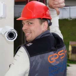 Instalator w czerwonym kasku, z uśmiechem spoglądający przez ramię, obok zamontowanej kamery monitoringu na ścianie budynku w Warszawie. Na kamizelce logo firmy GPS.