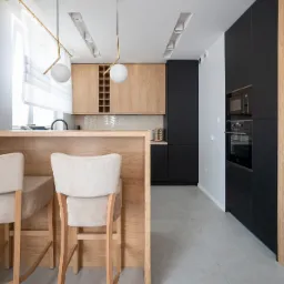 Jasna kuchnia z drewnianymi szafkami, czarnymi elementami i dwoma krzesłami barowymi z beżową tapicerką, oświetlona lampami wiszącymi z białymi kulami.