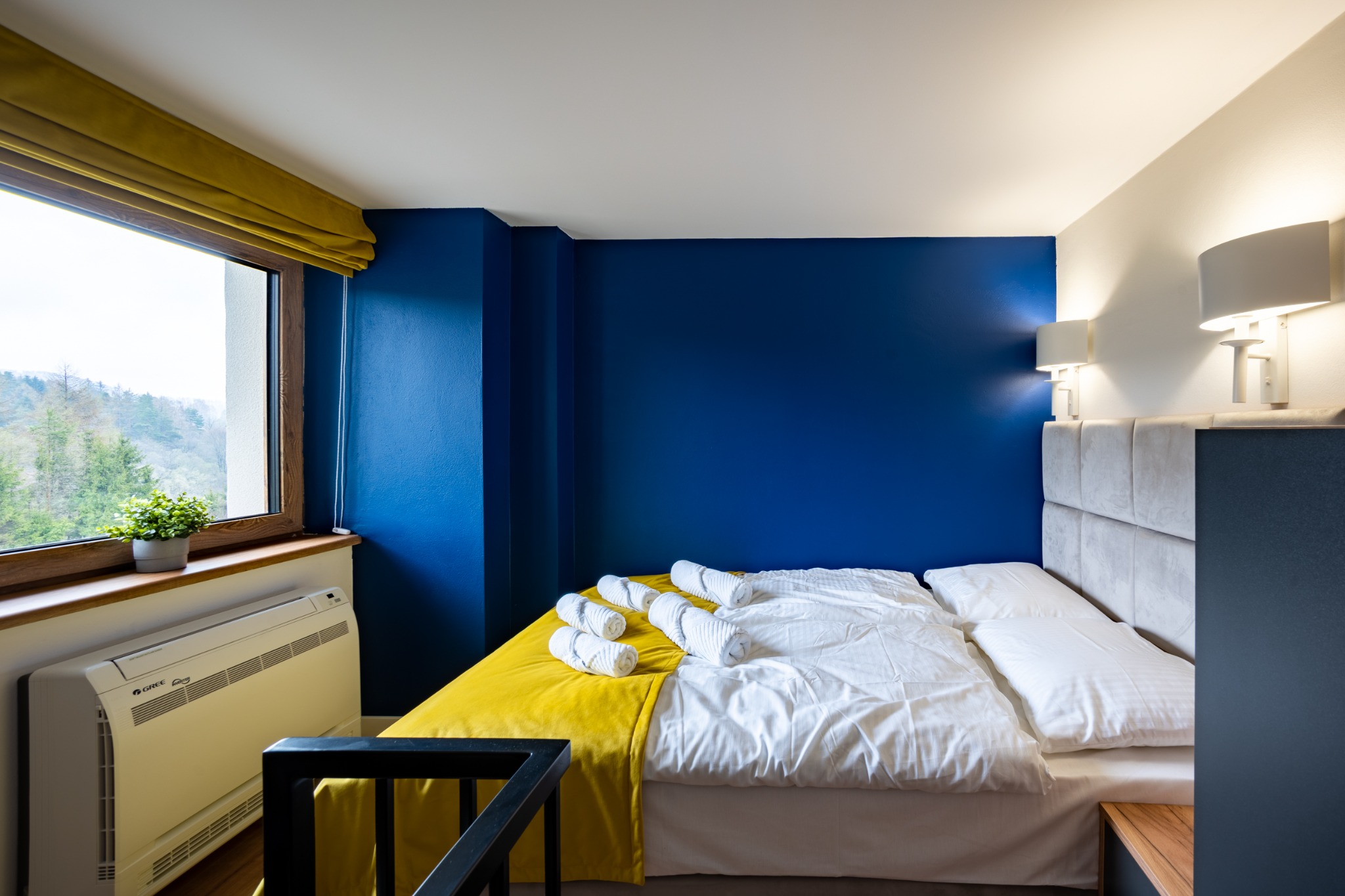 Pokój hotelowy z intensywnie niebieską ścianą, żółtą narzutą i zwiniętymi białymi ręcznikami na łóżku, widok na zewnątrz przez okno z rośliną doniczkową.