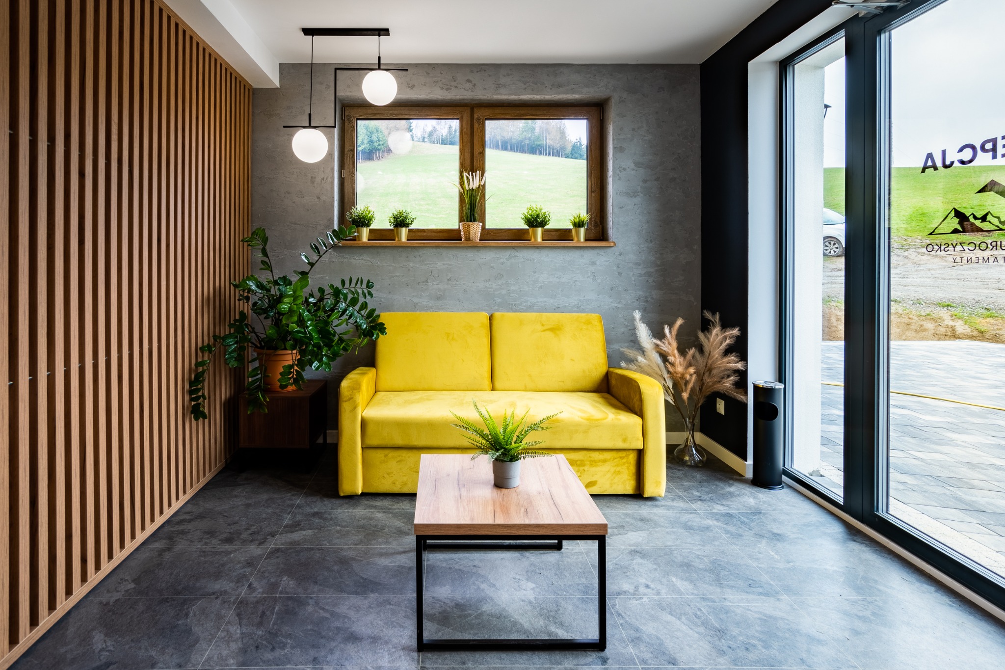 Jasnożółta sofa w nowoczesnym salonie z drewnianą ścianą, szarą podłogą i widokiem na zieleń za oknem.