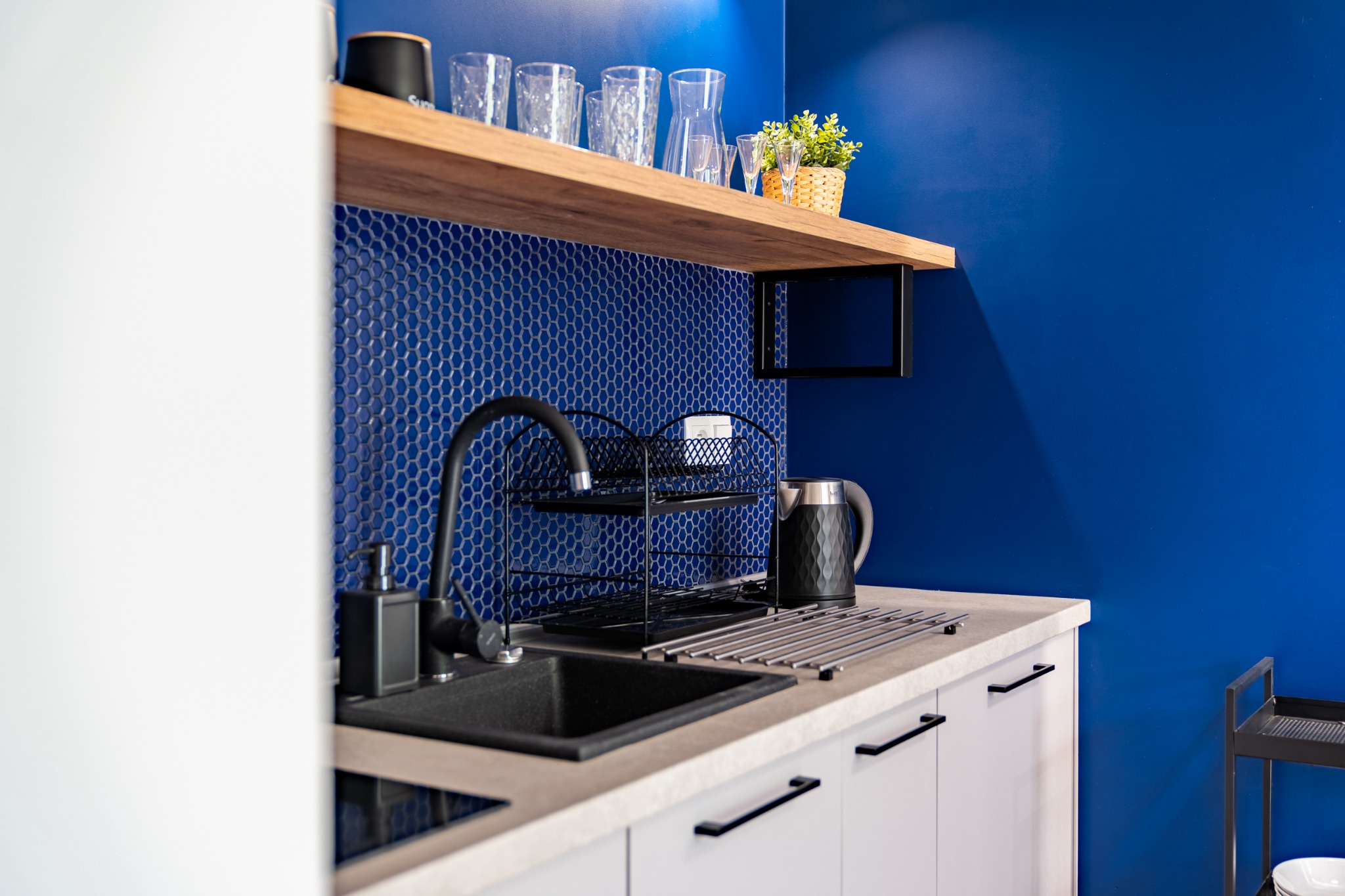 Nowoczesna kuchnia z niebieskimi, heksagonalnymi płytkami, drewnianą półką z dekoracjami, czarnym zlewem i armaturą oraz białymi szafkami z czarnymi uchwytami.
