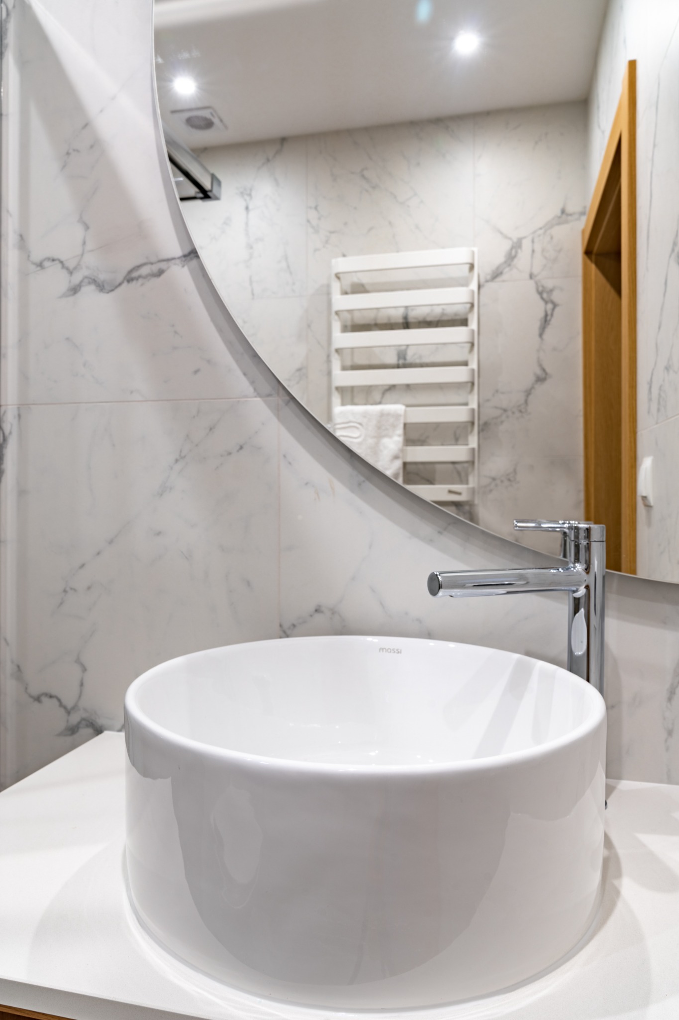 Biała, okrągła umywalka nablatowa marki Mossi z chromowaną baterią na tle marmurowej ściany i okrągłego lustra w nowoczesnej łazience.