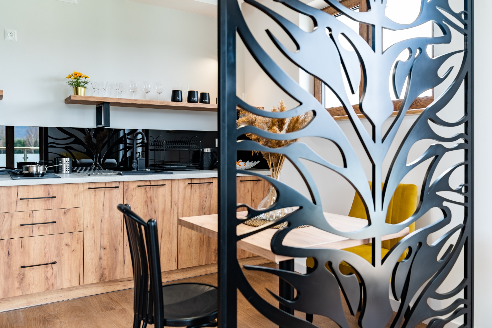 Nowoczesna kuchnia z drewnianymi frontami szafek, widziana przez dekoracyjny, ażurowy panel działowy w kolorze czarnym.