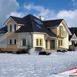 Dom jednorodzinny z ciemną dachówką karpiówką, widoczne okna dachowe, elewacja w odcieniach beżu i żółci, zimowy krajobraz z pokrywą śnieżną.