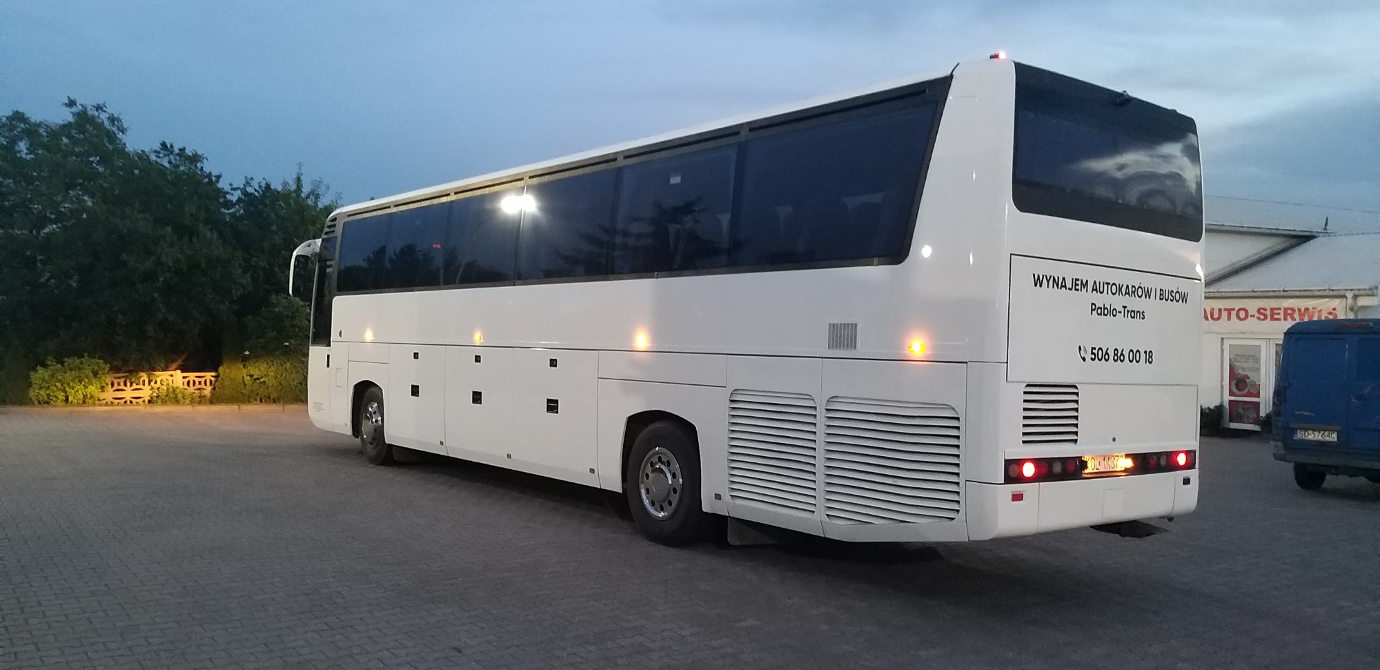 Biały autokar firmy Pablo-Trans z numerem telefonu na tylnej ścianie, zaparkowany na brukowanym placu w szarą pogodę.