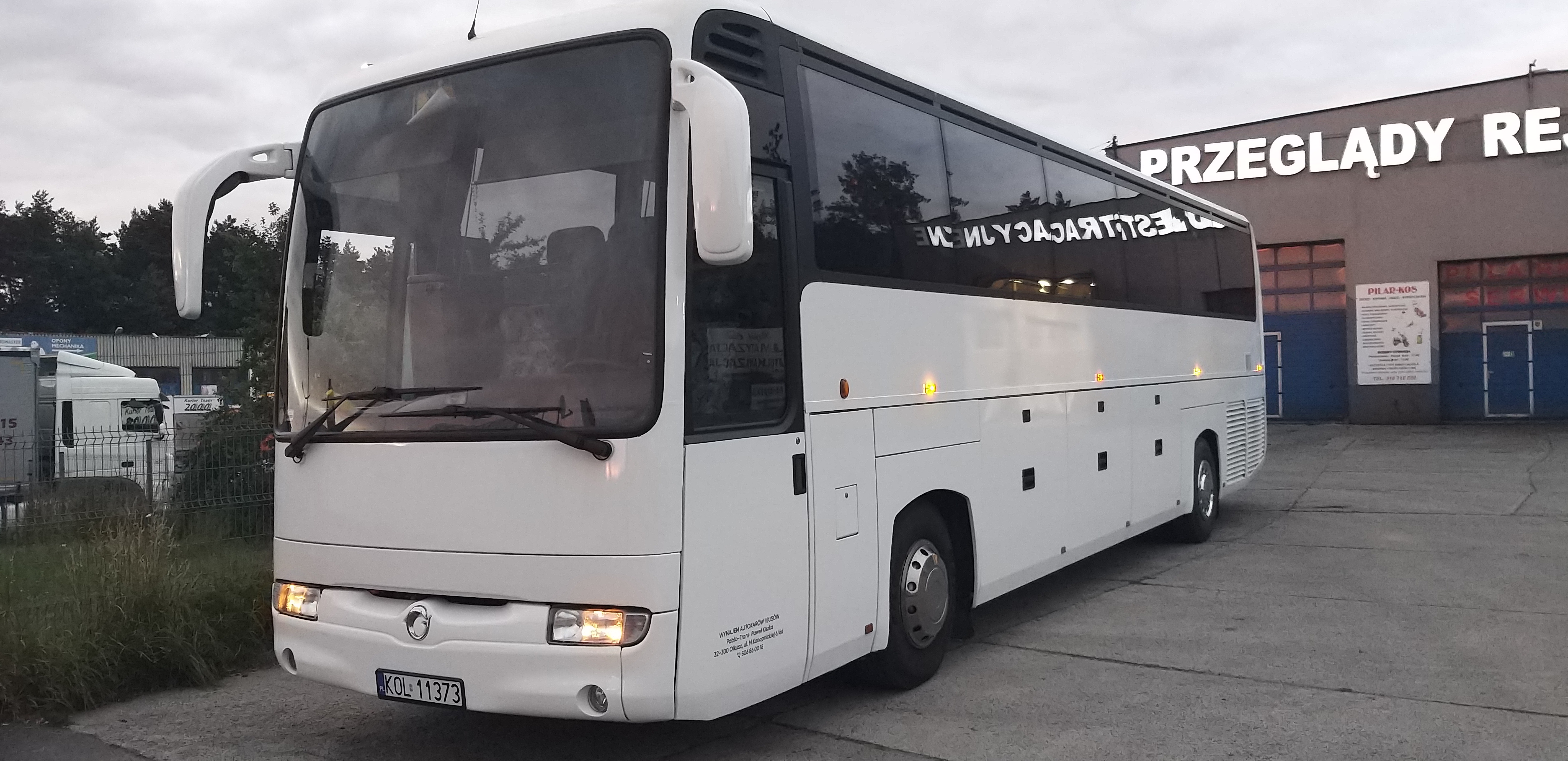 Biały autokar turystyczny zaparkowany przed budynkiem stacji kontroli pojazdów; widoczne odbicia w oknach.