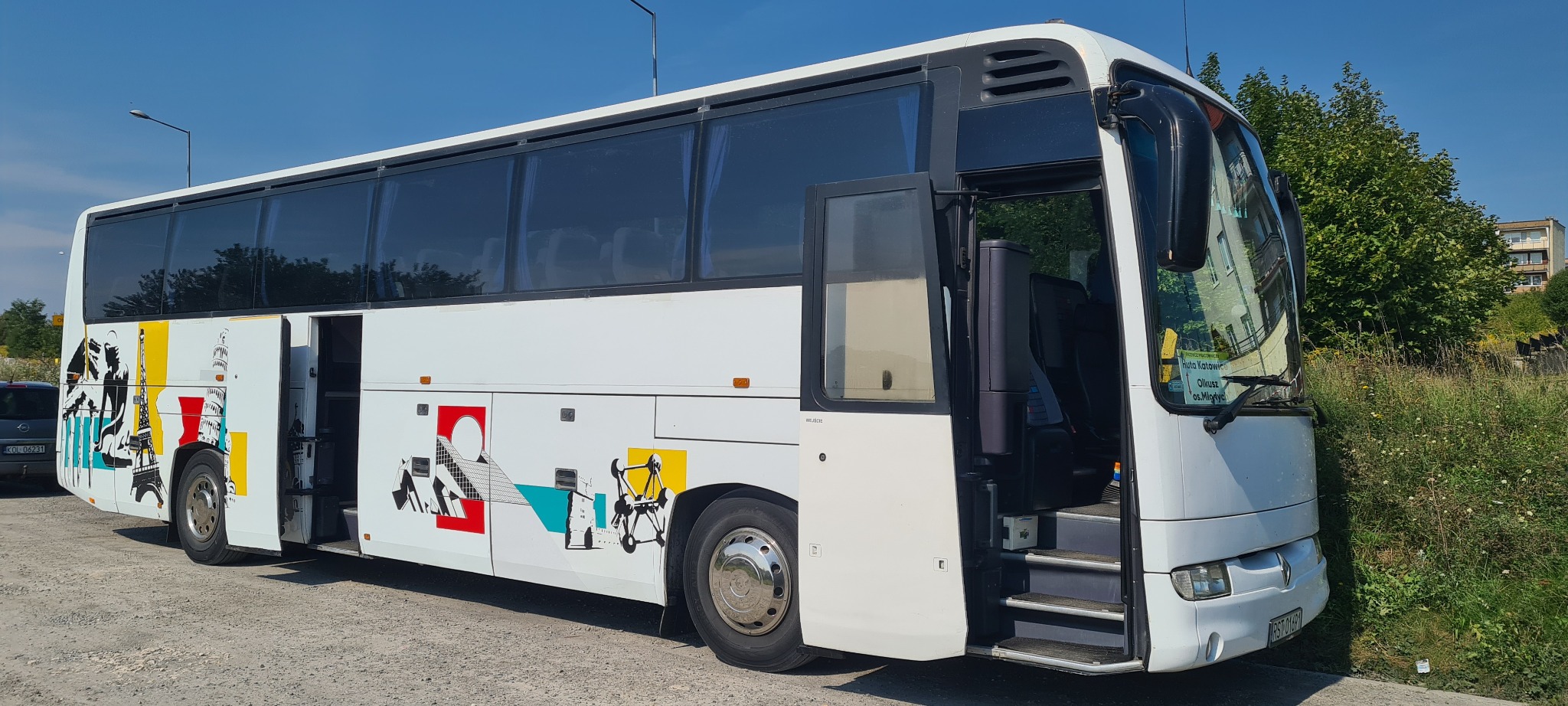 Biały autokar z otwartymi drzwiami, ozdobiony kolorowymi grafikami przedstawiającymi znane budowle i symbole, zaparkowany na żwirowym parkingu w słoneczny dzień.