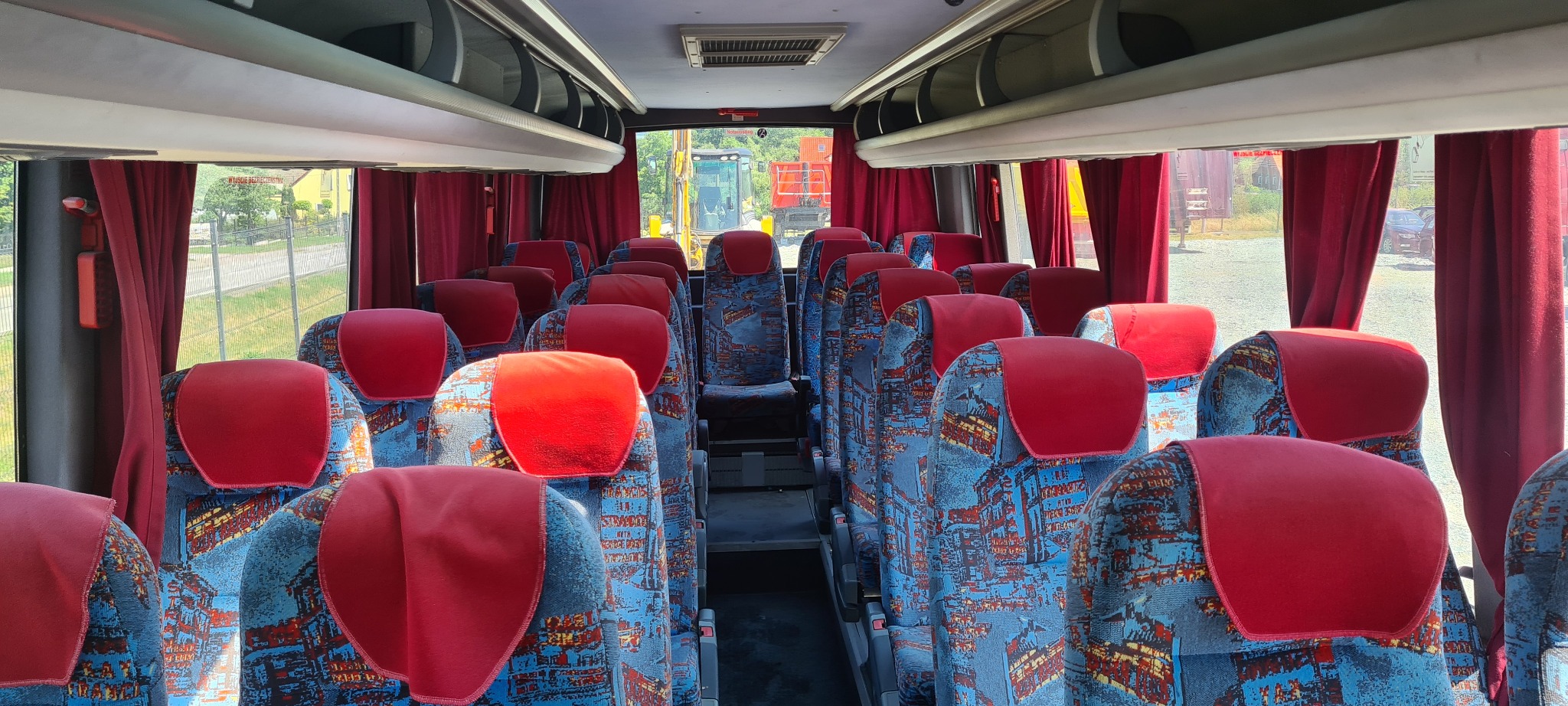 Wnętrze autobusu z rzędami foteli obitych wzorzystą tkaniną w odcieniach niebieskiego i czerwonego, czerwone zagłówki, zasłonięte okna z widokiem na zewnątrz.