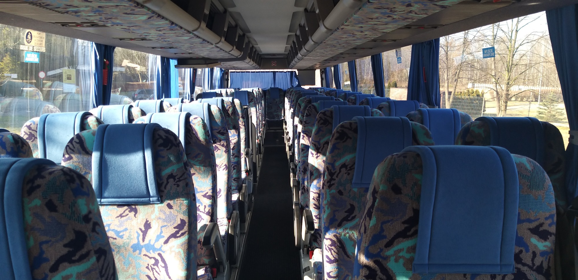 Wnętrze autobusu turystycznego z rzędami niebieskich foteli z wzorzystą tapicerką, widok wzdłuż korytarza w kierunku przodu pojazdu, zasłony przy oknach.