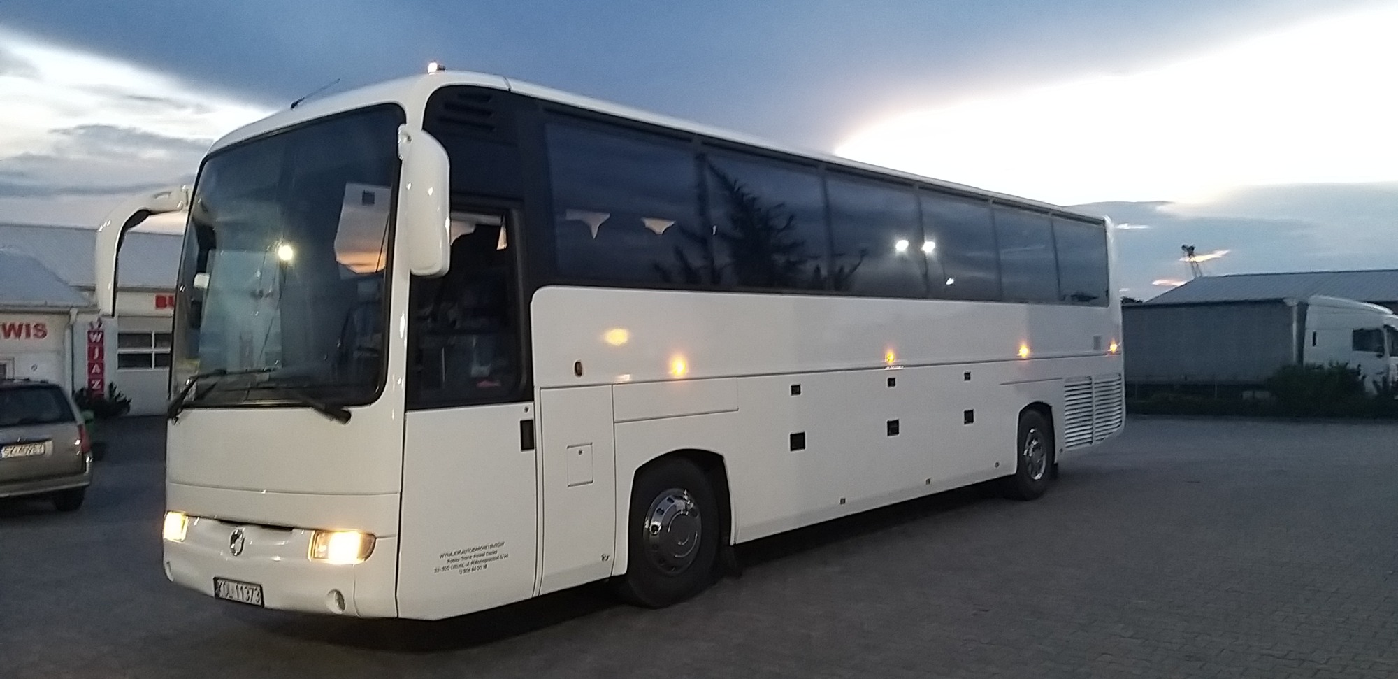 Biały autokar turystyczny na parkingu o zmierzchu, widoczne zaparkowane samochody i budynki w tle.