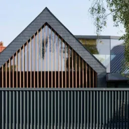 Nowoczesny dom z szarym dachem i drewnianymi elementami fasady, widoczny zza metalowego, pionowego ogrodzenia w odcieniach szarości i zieleni.