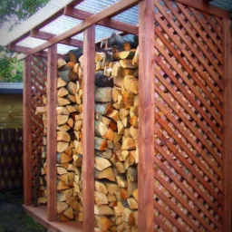 Drewniana konstrukcja z kratownicą i przezroczystym dachem, wypełniona pociętym drewnem. Widoczne elementy zaimpregnowanego drewna i betonowe podstawy.