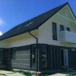 Nowoczesny dom jednorodzinny z czarnym dachem, białymi ścianami i grafitową elewacją, widoczne okna z czarnymi ramami i mały balkon z drewnianą balustradą.