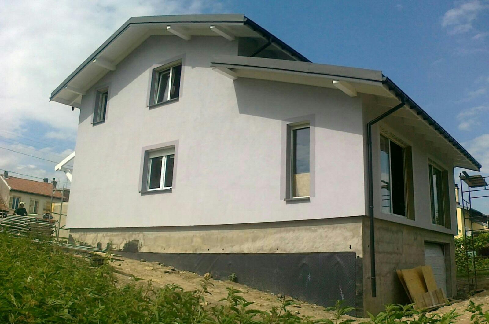 Dwukondygnacyjny dom z jasną elewacją, szarym dachem i białymi elementami wykończeniowymi, widoczny z poziomu gruntu, z fragmentem rusztowania i zielenią w pierwszym planie.