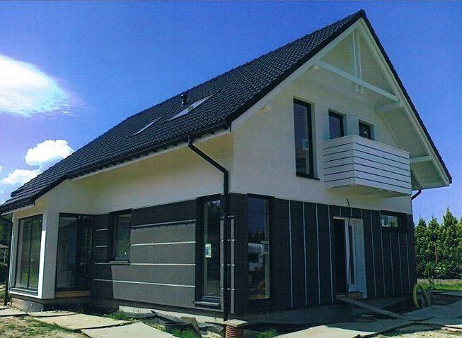 Nowoczesny dom jednorodzinny z czarnym dachem, białymi ścianami i grafitową elewacją, widoczne okna z czarnymi ramami i mały balkon z drewnianą balustradą.