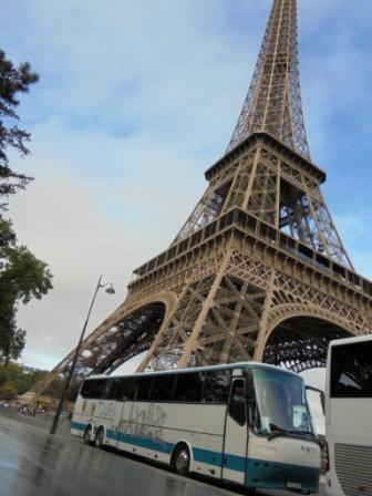 Autokar turystyczny na tle wieży Eiffla w Paryżu, pochmurne niebo. Autobus w barwach biało-turkusowych, z widocznym fragmentem drugiego pojazdu.