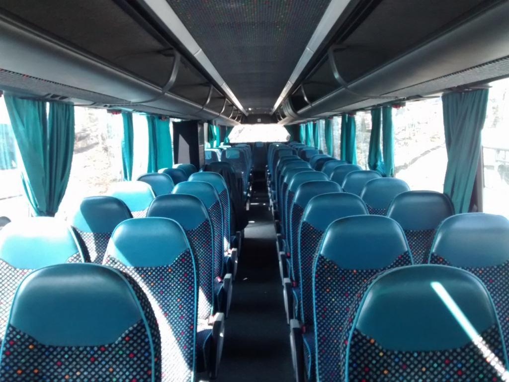 Wnętrze autobusu z rzędami niebieskich foteli z wzorem w kropki, zielone zasłonki przy oknach, widok z przodu pojazdu, oświetlenie sufitowe.