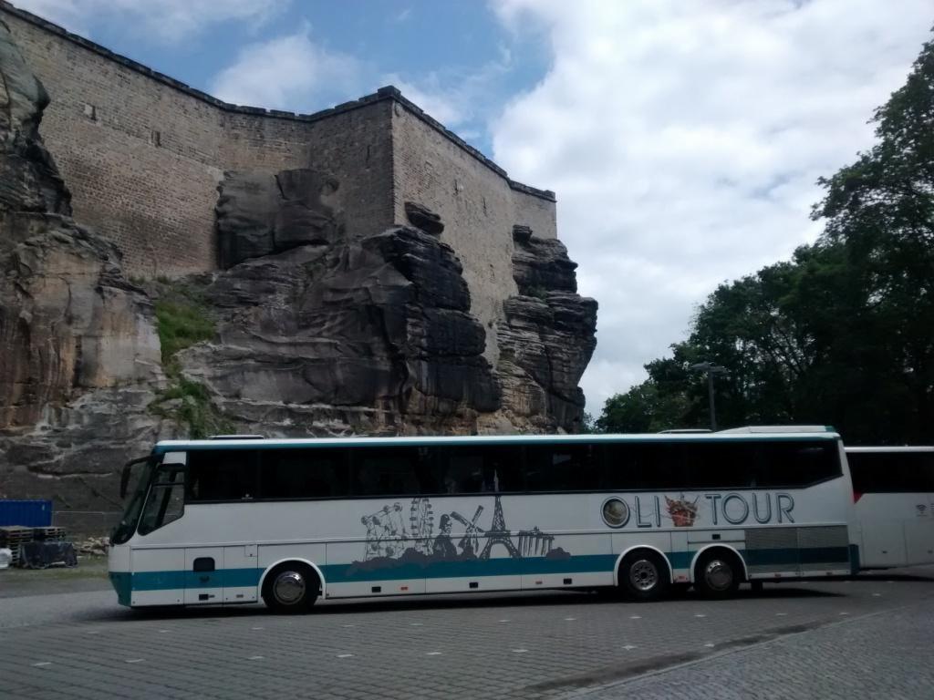 Autokar turystyczny z grafiką panoramy miasta na tle skalnej ściany i zamku, zaparkowany na brukowanym placu w pochmurny dzień. Widoczny fragment drugiego autokaru.
