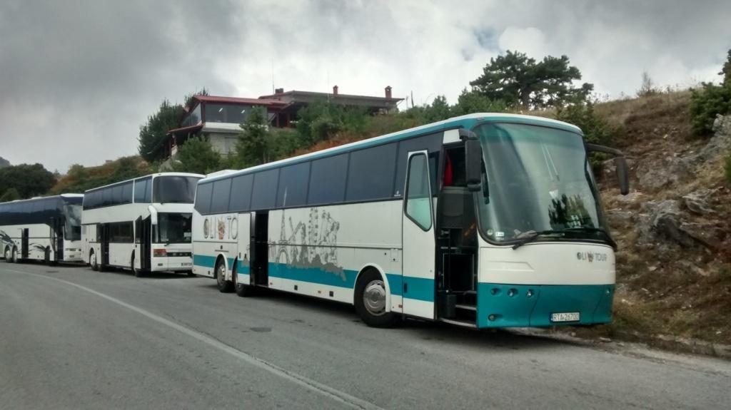 Flota autokarów turystycznych z otwartymi drzwiami, zaparkowana na poboczu drogi, z budynkiem w tle, przy pochmurnym niebie.