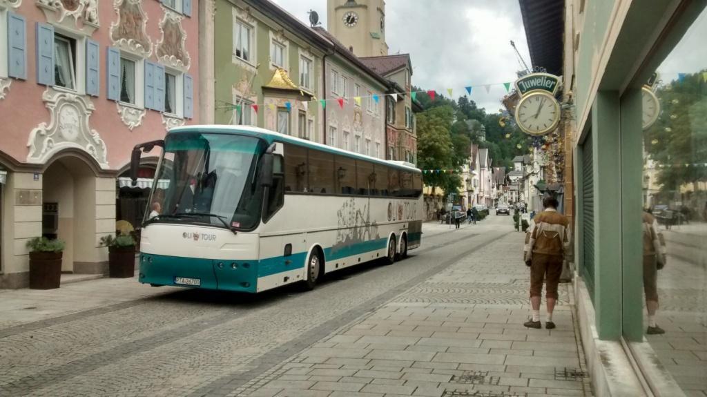 Autokar turystyczny OLI TOUR na tle malowniczego miasteczka z flagami i zegarem Juwelier. Osoba w tradycyjnym stroju. Refleksja w witrynie sklepowej.