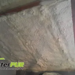 Izolacja poddasza wykonana metodą natrysku, widoczne nierówności i struktura piany, drewniana belka konstrukcyjna i fragment ściany z pustaków.