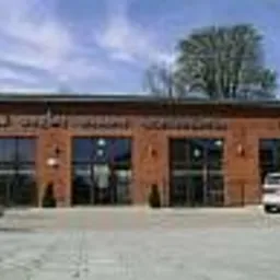 Elewacja budynku z cegły z dużymi, przeszklonymi drzwiami przesuwnymi, widoczne zaparkowane samochody i drzewo w tle, słoneczny dzień.