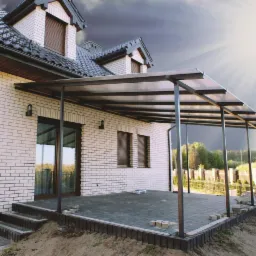 Metalowa pergola przy białym domu z cegły, z widocznym zadaszeniem i częściowo wykończonym tarasem z kostki. Niebo pochmurne, z promieniami słońca.