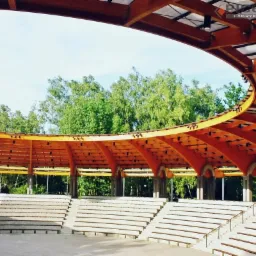 Drewniana konstrukcja zadaszenia amfiteatru z widownią schodkową, otoczona zielenią drzew. Widoczne elementy oświetlenia i logo producenta.