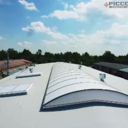 Biały dach przemysłowy z świetlikami kopułowymi i kominkami wentylacyjnymi. Widok z góry, błękitne niebo z chmurami w tle. Logo producenta w prawym górnym rogu.