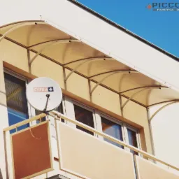 Lekka, łukowa osłona nad balkonem z widoczną anteną satelitarną. Jasna kolorystyka budynku i błękit nieba w tle. Widok z dołu.