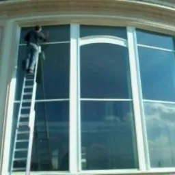 Mężczyzna na drabinie myje duże, łukowe okno w jasnej elewacji budynku. Widoczne odbicie nieba w szybach, co sugeruje czystość. Praca na wysokości.