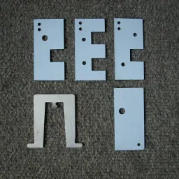 Pięć metalowych elementów o różnych kształtach (E, prostokąt, odwrócone U) z wywierconymi otworami, ułożonych na szarym, teksturowanym tle.
