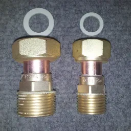 Dwa mosiężne złączki z gwintem zewnętrznym i miedzianym elementem, leżące na szarym dywanie, z dwiema białymi uszczelkami u góry. Jedna złączka jest większa i oznaczona '18x3/4 Rotgu', druga...