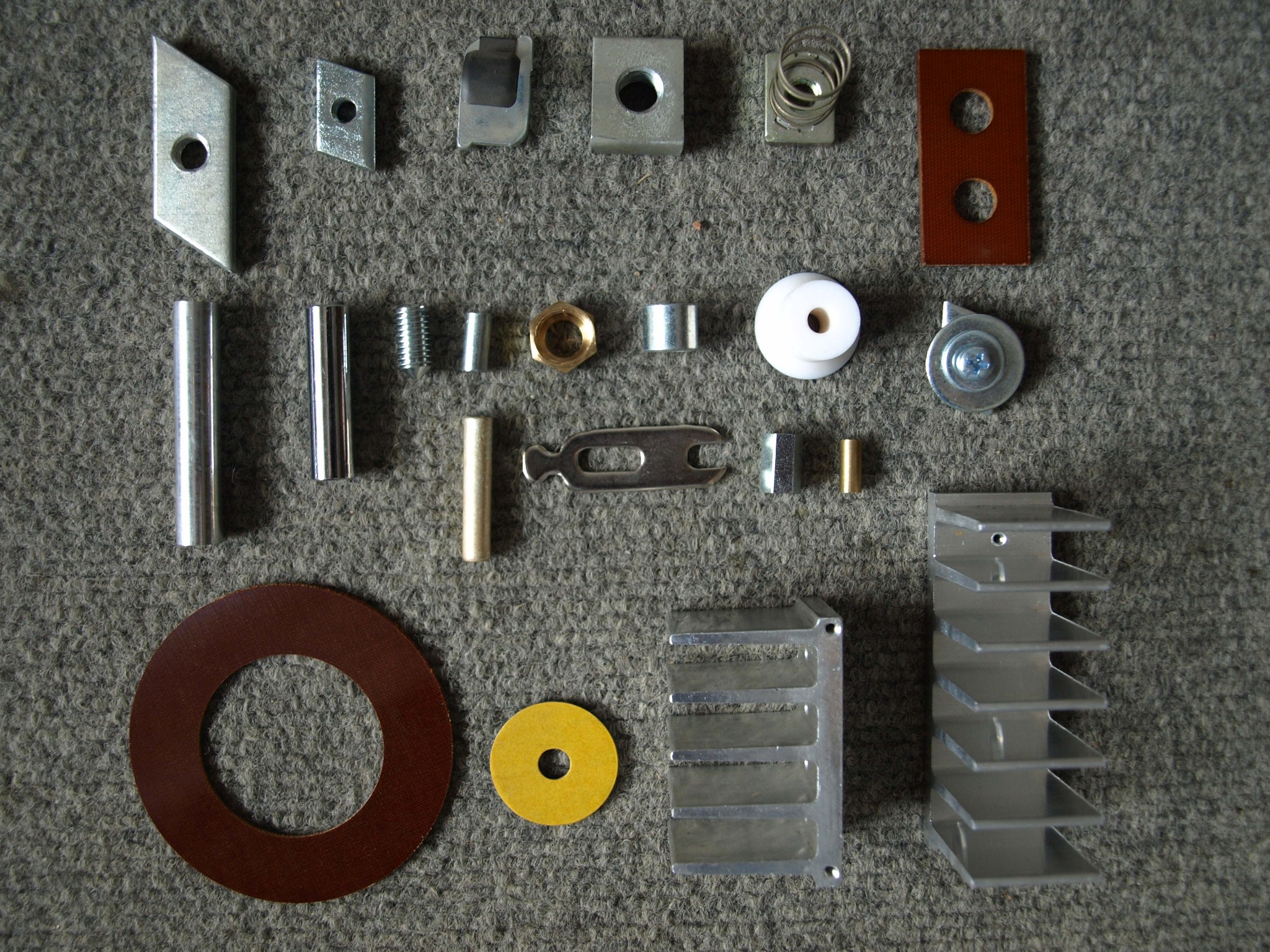 Zestaw różnorodnych, metalowych i niemetalowych elementów maszynowych, w tym śruby, nakrętki, podkładki, tuleje, sprężyny i radiatory, ułożonych na szarym, teksturowanym tle.