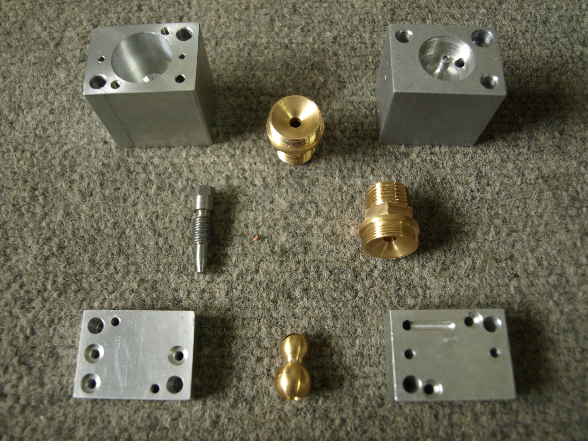 Precyzyjnie obrobione metalowe elementy: dwa aluminiowe bloki z otworami, dwa mosiężne elementy z gwintem zewnętrznym i wewnętrznym, aluminiowa płytka z otworami oraz mosiężna figurka w kształcie...