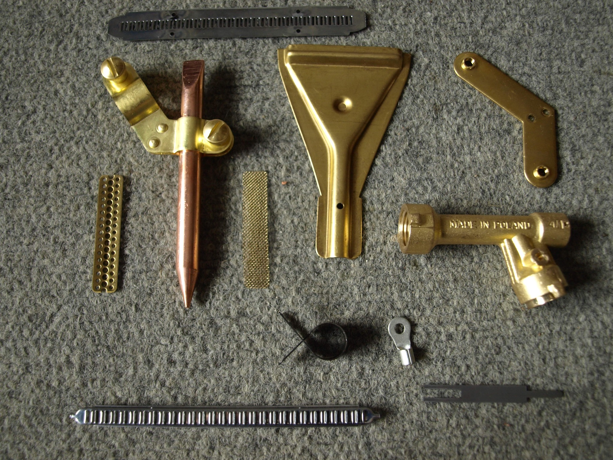 Zestaw różnorodnych, metalowych elementów i akcesoriów lutowniczych, w tym dysze, uchwyty, sprężyny i złączki, ułożonych na szarym, teksturowanym tle.