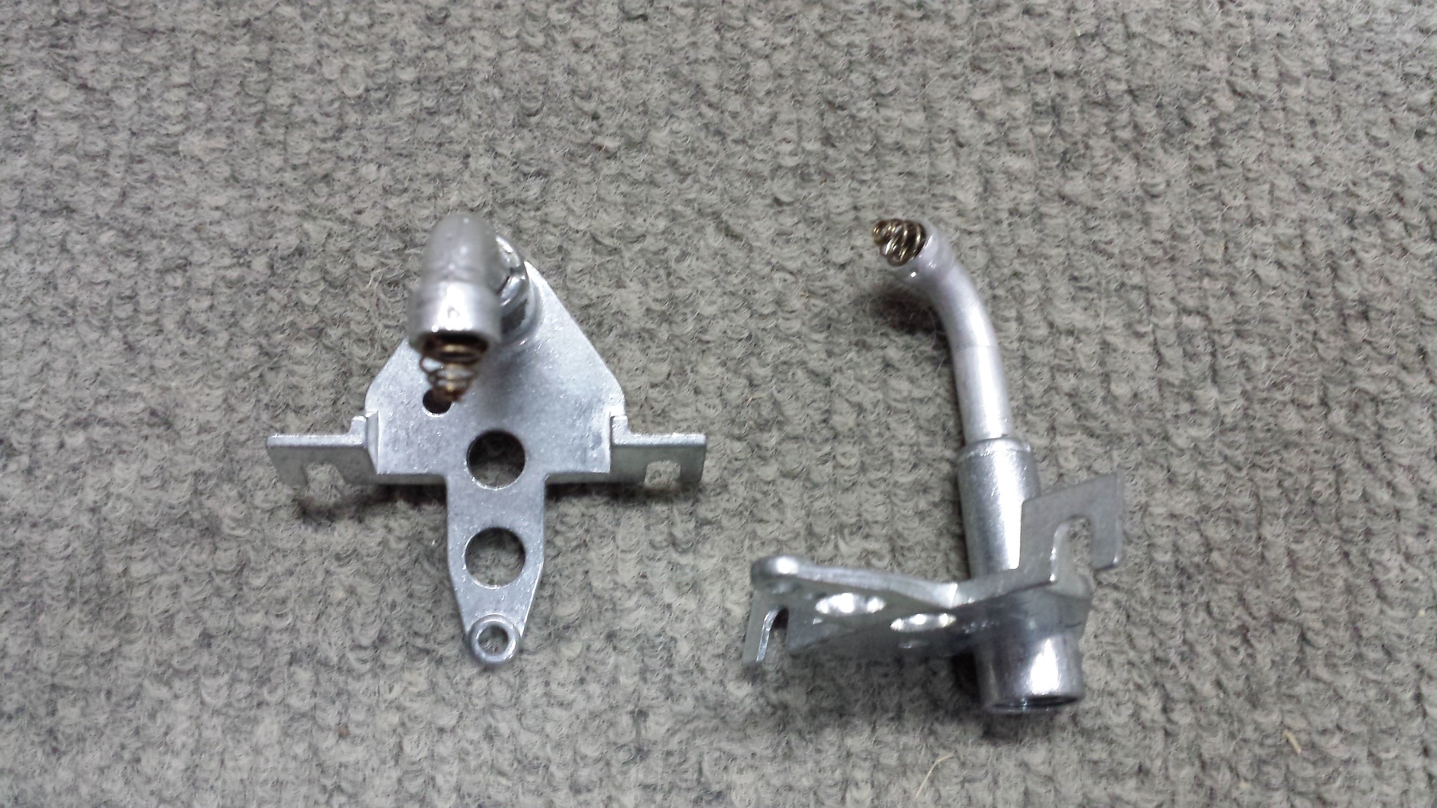 Dwa srebrne, metalowe elementy obrobione maszynowo, leżące na szorstkim, szarym tle. Jeden element ma kształt trójkąta z otworami i sprężyną, drugi ma zakrzywioną rurkę i podstawę z otworami.