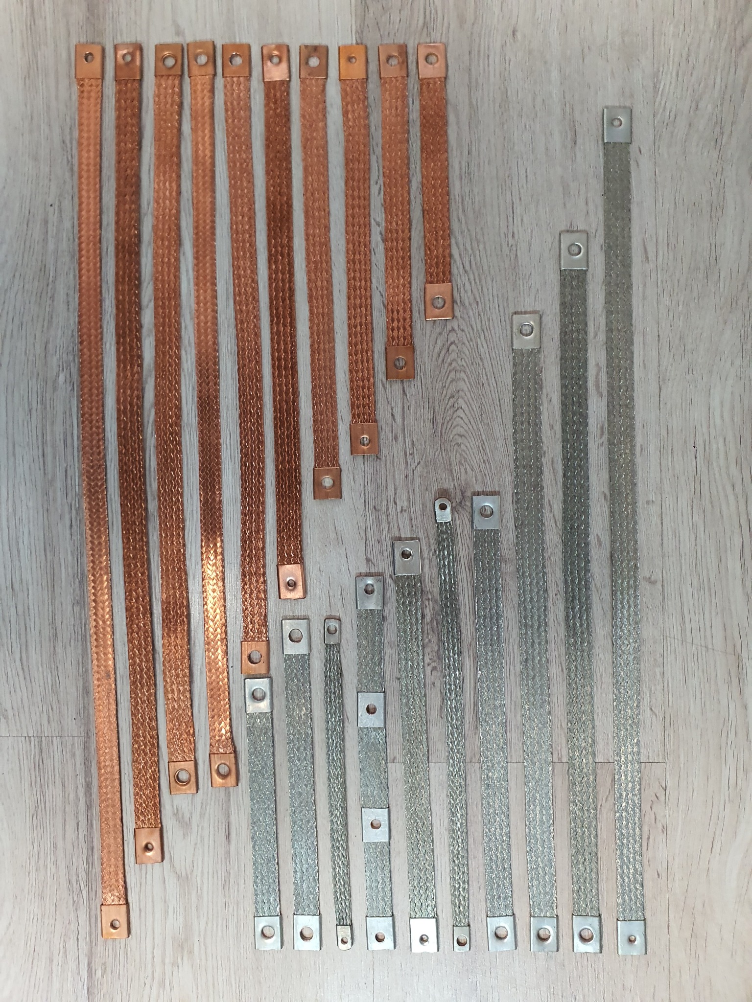 Zestaw plecionych, miedzianych i posrebrzanych przewodów uziemiających o różnej długości, zakończonych kwadratowymi końcówkami z otworami montażowymi, ułożonych na drewnianym podłożu.