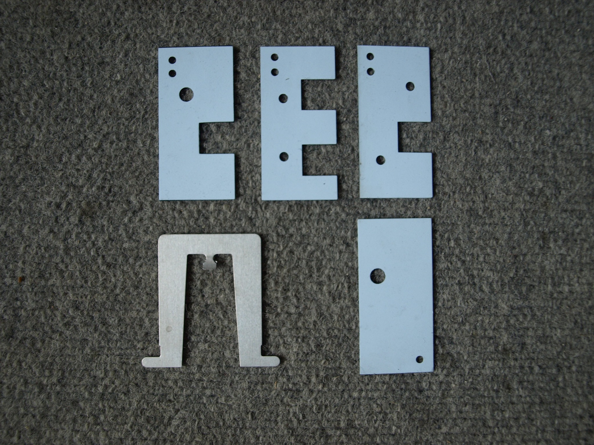 Pięć metalowych elementów o różnych kształtach (E, prostokąt, odwrócone U) z wywierconymi otworami, ułożonych na szarym, teksturowanym tle.