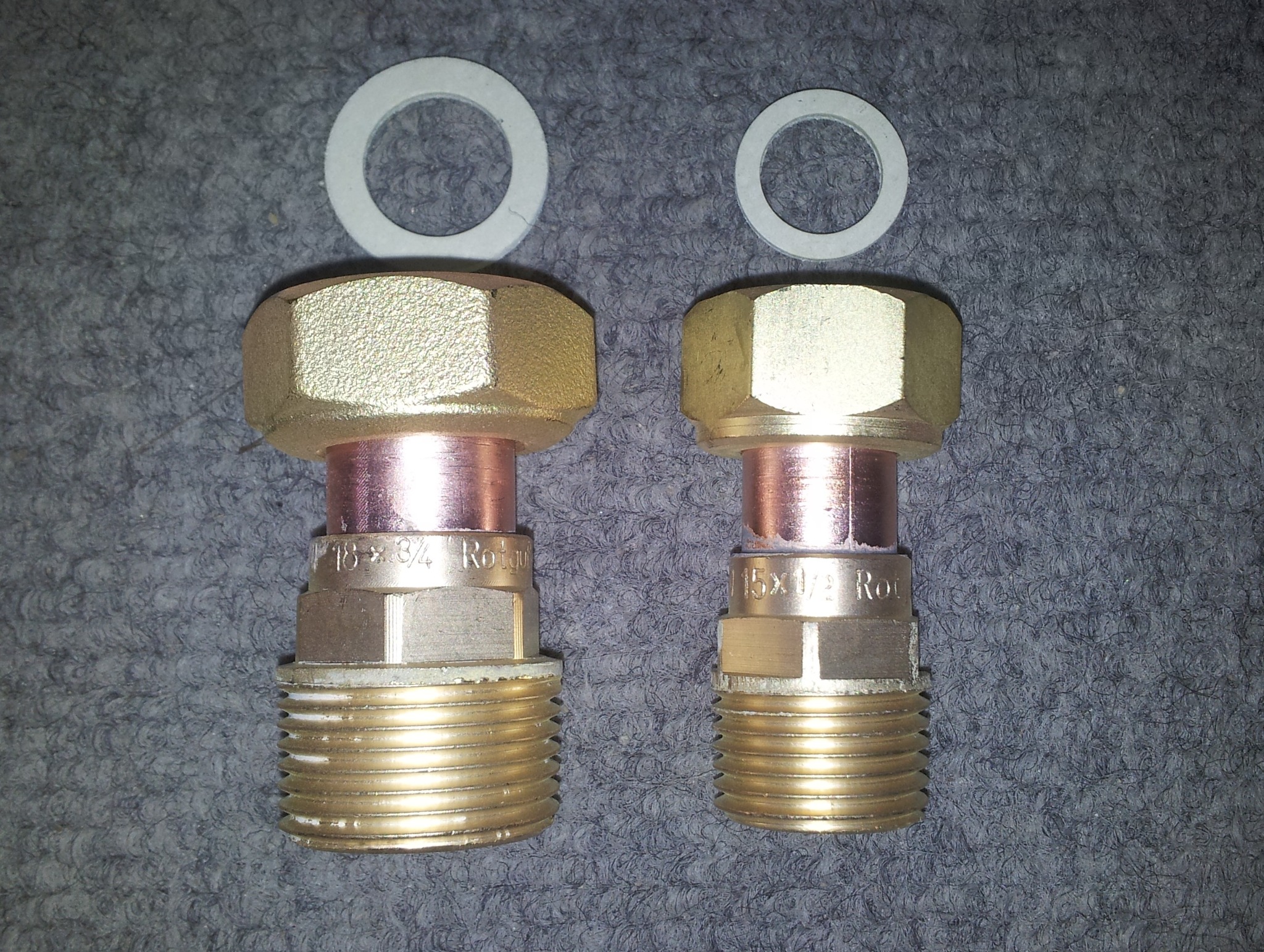 Dwa mosiężne złączki z gwintem zewnętrznym i miedzianym elementem, leżące na szarym dywanie, z dwiema białymi uszczelkami u góry. Jedna złączka jest większa i oznaczona '18x3/4 Rotgu', druga...