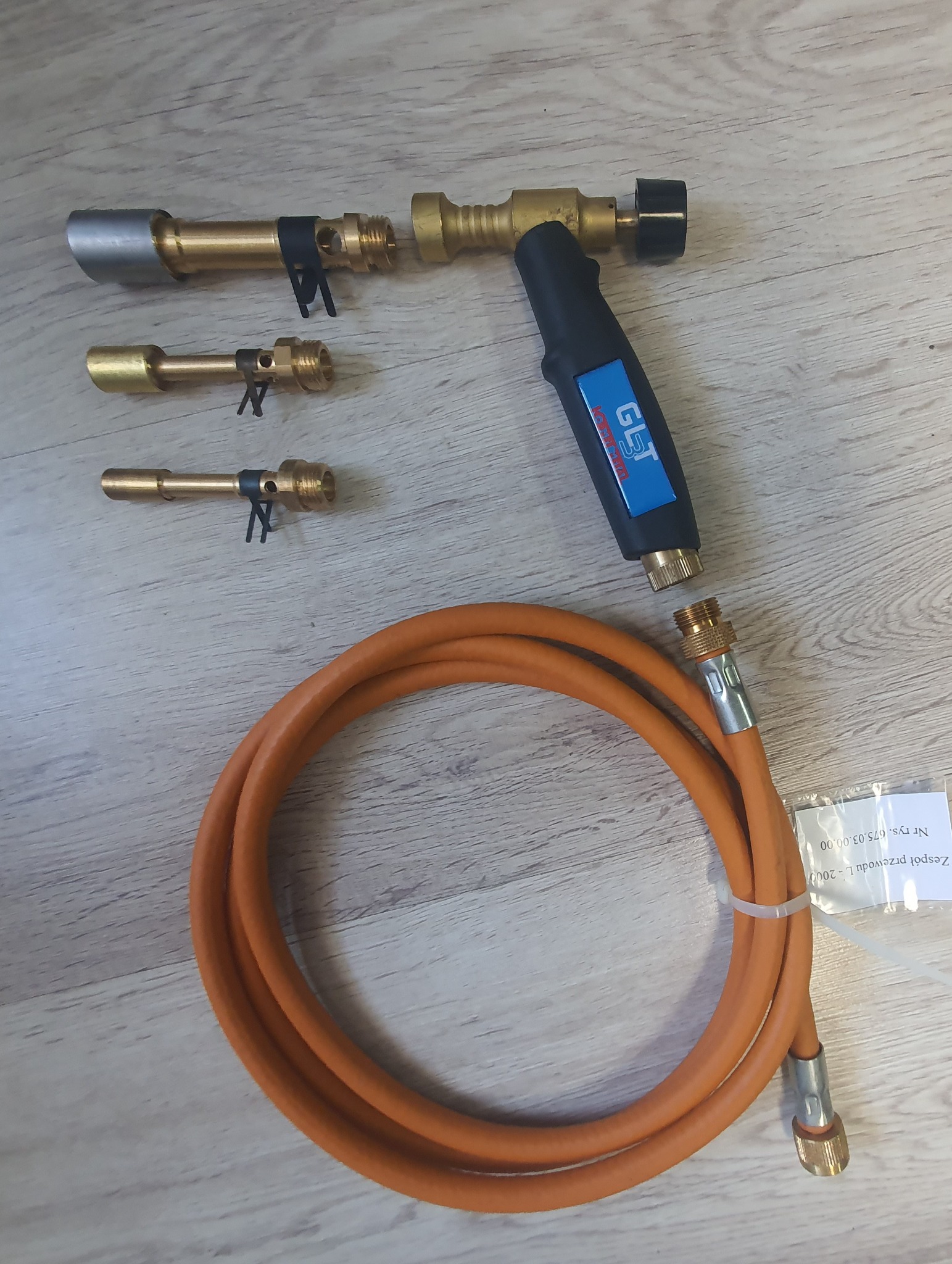 Zestaw palników gazowych z dyszami o różnych średnicach i pomarańczowym wężem gazowym na jasnym drewnopodobnym podłożu.