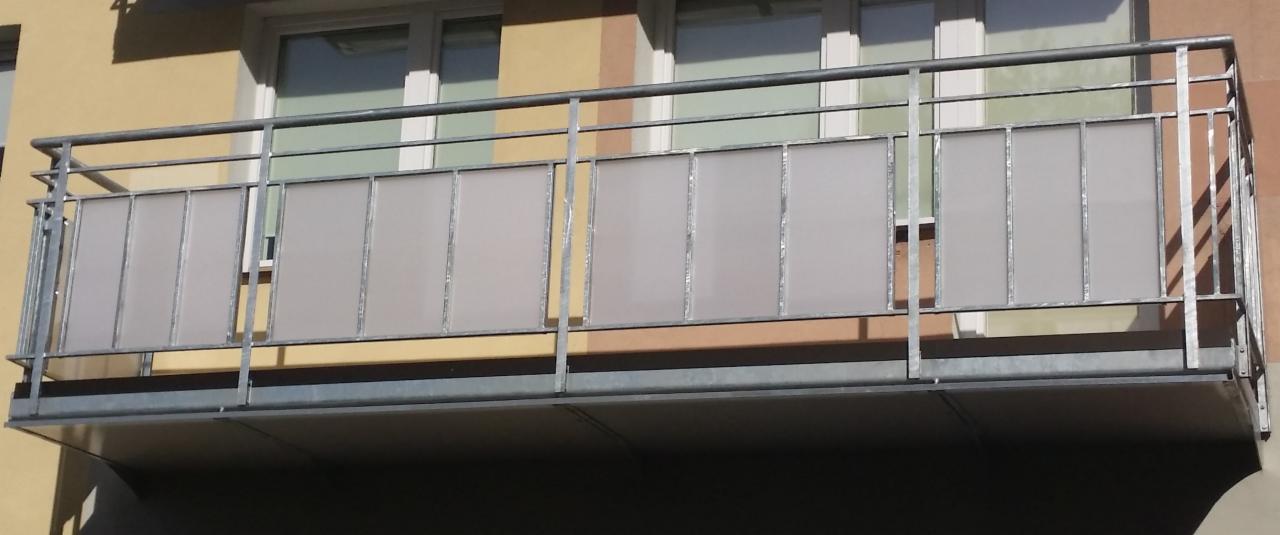 Balkon z metalową balustradą wypełnioną matowymi panelami, widoczny z dołu, na tle elewacji budynku z oknami.